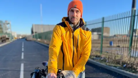 Dan is wearing a yellow jacket as he straddles his bike on a road. He has an orange hat on.