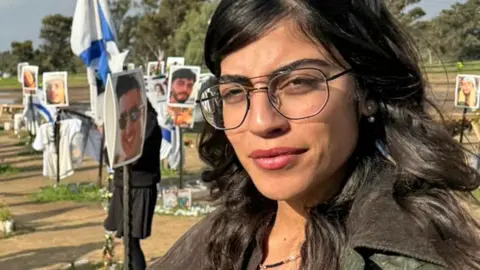 Oren Rosenfeld / BBC Michal Ohana stands with long dark hair and large glasses and wearing pink lipstick, in front of a memorial for those who died on October 7 at the Nova Festival, with pictures of people who were killed, Israeli flags, flowers and other feed.