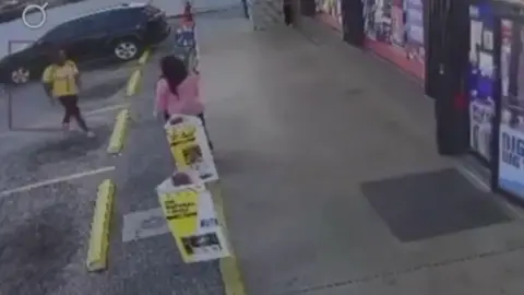 An image from surveillance video shows a woman in a pink blouse standing outside a convenience store as a man in black pants and a yello shirt approaches her from the left