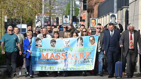 People walking carrying a large banner reading: "Springhill-Whiterock Massacre Belfast's Bloody Sunday Time for Truth" with pictures of the five deceased. 