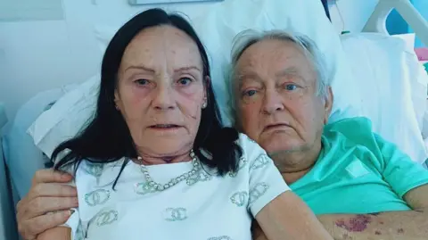 Supplied Bill Trotter, a man in his 70s, is lying in a hospital bed next to his wife who is a similar age. He has short, grey hair and she has shoulder-length black hair. They both look frightened. He's wearing a green hospital T shirt, she has a white top and is wearing a necklace of pale green beads or pearls. Behind are some white hospital pillows.