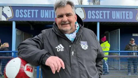 Prostate Cancer UK The image shows a football ground, with a man standing in front of a stand displaying the text Ossett United along the top fascia. The man is pitchside, with railings, signage and spectators visible. He is wearing a dark Ossett United jacket featuring the club’s crest and the initials “DB” on the chest.