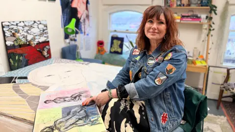 A woman with brown-red hair smiles into the camera. She is wearing a blue denim jacket covered in fabric patches and a black and white dress. She is sitting at a table with a sketch book. Decorative artwork and lamps are visible in the background.
