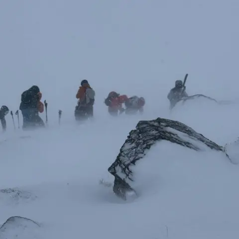 SAIS Southern Cairngorms The ski tourers stand in drifting snow near a rocky outcrop.