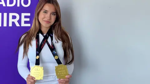 Victoria standing in front of a banner. She has long brown hair and is wearing a long-sleeved white top and is holding her two gold medals