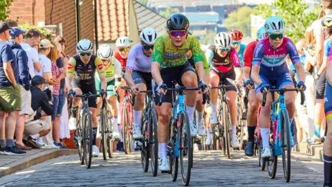 A group of cyclists wearing colourful cycling kit riding towards the camera on a narrow cobbled street. Their faces show the pain and effort of climbing up a steep hill. Spectators line the paths next to the road, with a view looking downhill towards the buildings of a city centre in the background.