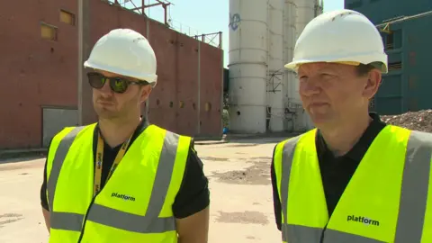Neil Rustage (left) and John Laverick (right) on the former Unilever site. They are both wearing hard hats and high visability vests. The former industrial buildings are in the background.