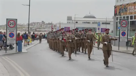 Soldiers in khaki uniforms march in rows along the seafront at Bridlington. Crowds have gathered to watch them.