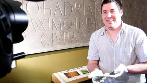 British Museum Portable Antiquities Scheme Picture of Peter Reavill wearing white gloves and holding polished stones in his hand, standing against a backdrop of a large Latin-inscribed stone placard on the wall behind him. He is looking at a film camera which is partially in shot.