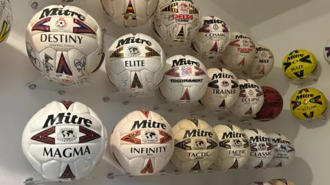 A large amount of Mitre footballs mounted and displayed above a staircase on a plain white wall. Each ball has a different Mitre model and design. The majority are white coloured but there also yellow and red ones. Some also have signatures on them.