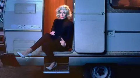 Michelle Collins in a Marilyn Monroe-styled blonde curly wig and black polo neck and leggings with white stiletto shoes sits on the steps of a caravan