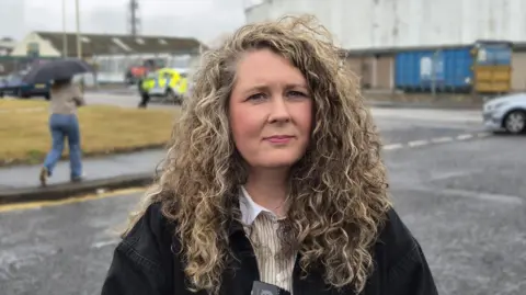 Councillor Lois Speed has curly blonde hair and is wearing a black jacket. She is standing outside and there is a police car behind her