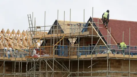 Two builders working on the construction of new homes. They are both on the roof in high-vis jackets and dark clothing. You can see scaffolding, a roof being built, and wooden timber. 