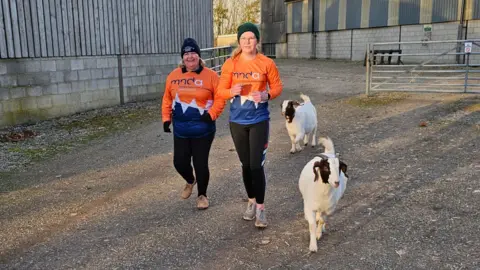 Megan Wilson Two women, in orange MND t-shirts, run alongside two goats on a farm.