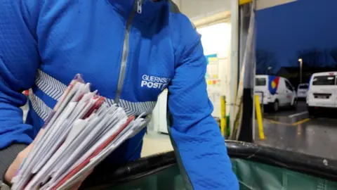 Photo of someone's torso with a Guernsey Post branded blue zip-up jacket. They are holding a stack of letters in their hand.