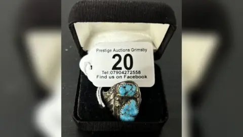 Prestige Auctions A silver ring in a black ring box with a white label on it stating "Prestige Auctions Grimsby". The ring is silver with indentations and turquoise stones. 