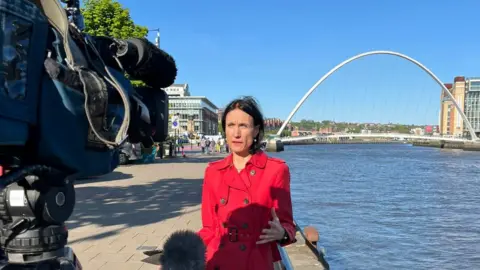 Fiona Trott standing outside Newcastle Crown Court beside the River Tyne with the Gateshead Millennium Bridge behind her. She is wearing a red jacket and is facing a camera and there is also a microphone in shot
