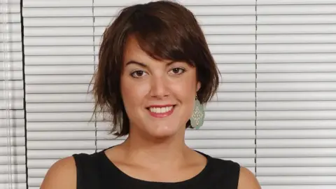 Yasmina Siadatan is sat on a sofa in front of closed white blinds. She has brown hair and is wearing green earrings and smiling for the picture. 