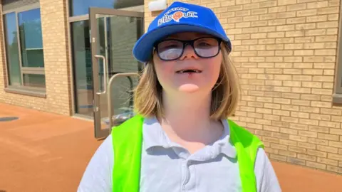Sam Read/BBC Lily, standing outside, wearing a white top, with a high-vis jacket, she has glasses on, is wearing a blue hat and has shoulder length blonde hair. A school building is behind her.