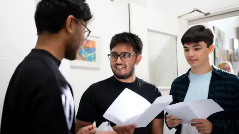 Getty Images Habib Matlib, Umair Abdullah and John Bose receive their A-level results at Ffynone House School in Swansea, Wales. Umair is in the centre and is wearing a black T-shirt and black glasses - he is holding a sheet of paper with his exam results on and is looking at Habib, who is out of focus and can only be seen side-on. John is on the right of the three, wearing a pale blue T-shirt under a very dark blue checked shirt - both he and Umair are smiling.