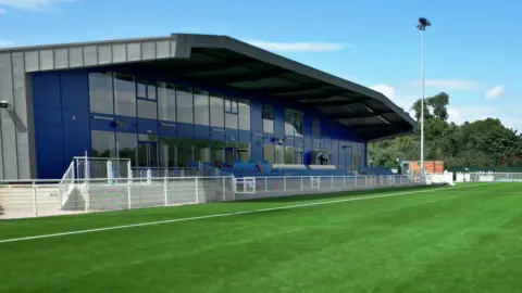 BBC Essex Sport A modern terrace building and clubhouse beside an immaculately green football pitch. There is blue sky above and some cloud.