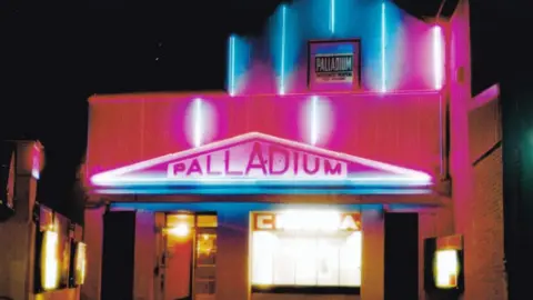 The cinema façade lit up at night in pink and bluish colours - with the word Palladium across the top of the front entrance in a white triangle. The cinema has an art deco look to it 