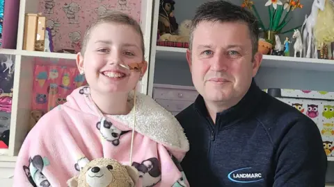 Family photo Megan and Matt. Megan has short blonde hair and a tube in her nose. She is wearing a pink fluffy hoodie covered in panda print. She is holding a beige teddy bear and smiling at the camera. Matt is sat next to her. He has dark short hair and is wearing a black jacket. He is smiling at the camera too. 