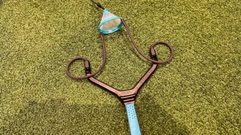 A catapult, with a turquoise handle and bronze frame and brown elastic, lies on a patch of green artificial turf.