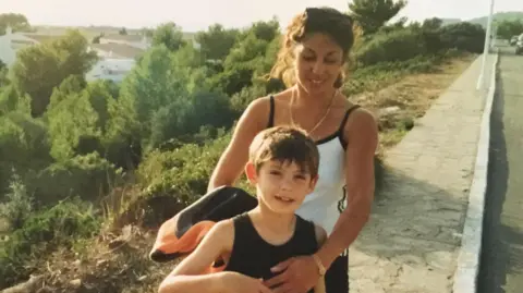 Mach 3 Studio Ltd Old film camera image of Olli and his mum on holiday standing on a path next to a road. He is a child and is wearing a black vest, whilst his mum is wearing a white vest and holding him.