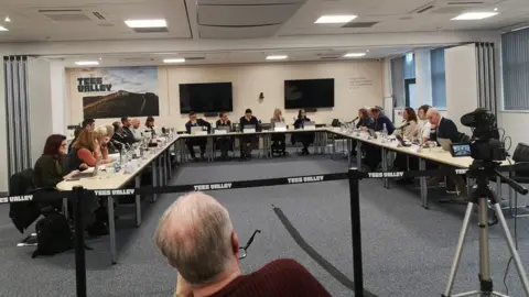A meeting of the TVCA. Councillors are sat at long tables arranged in a U-shape. Members of the public are sat on the other side of the room to watch the meeting. A sign reading Tees Valley hangs on the wall next to two TVs.