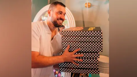 Unveil by Hollie A man with short brown hair and a beard and a white shirt is smiling while holding a pile of eight pizza boxes with a black and white chequered pattern on the sides.