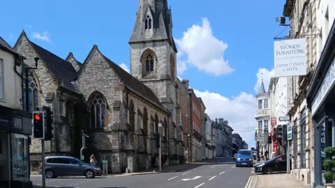 A view of Dorchester High Street, with a grey brick church and church tower, a traffic light to the left and shops along a street to the right. There are cars driving along the road.