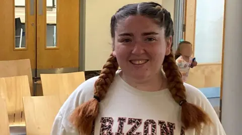 BBC Aimee is standing in a vaccination clinic's waiting room. She has long brown hair in plaited bunches and is wearing a white jumper as she smiles for the camera.