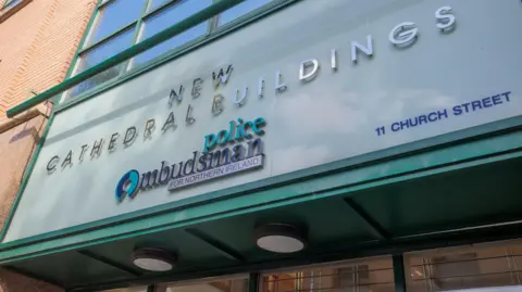 PA Media A brown brick building with a green sign with silver and blue writing with 'new cathedral buildings police ombudsman for Northern Ireland' written on it.