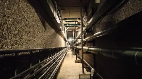 Simon Furber The networks underneath Guildford Cathedral. A series of pipes can be seen in the image on a long corridor. There is a light at the end.