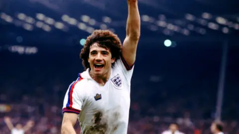 Kevin Keegan celebrates with his arm in the air after scoring a goal for England. He has short curly hair and is wearing a white England shirt with mud on the front.