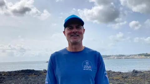 Un hombre con camisa y gorra azules sonríe mientras mira a la cámara. Detrás de él se ve la costa de Guernsey, así como las rocas de la playa, el mar y las nubes.