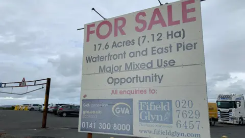 A for sale sign on Stranraer waterfront advertising the waterfront and east pier as a "major mixed use opportunity". The sign is in front of a car park with various vehicles in it under a cloudy sky.