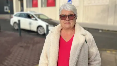 A woman with short white hair is wearing sun glasses, a white coat and red top