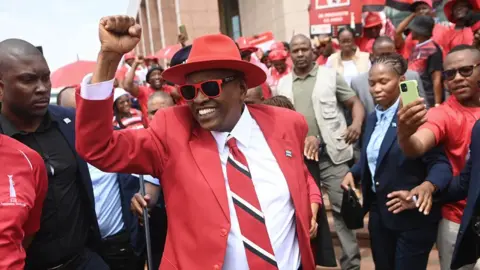 AFP Mokgweetsi Masisi dressed in a red suit and wearing a red hat and sunglasses holds his fist up as he is surrounded by supporters.