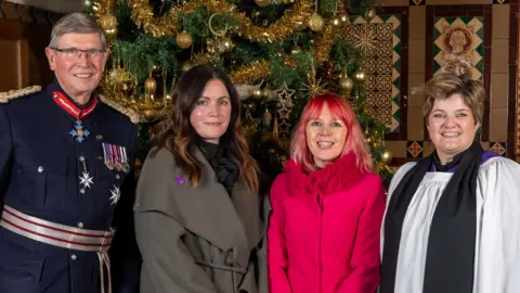 Four people stand in front of a Christmas tree decorated with gold tinsel and baubles. The man on the left is wearing a navy military uniform complete with medals on his left breast and chest. The lady to his left has long dark hair with a grey flowing coat. To her left is a lady with bright red hair, fading to pink at the tips. She is wearing a bright pink coat with a furry pink collar. To her left is a female priest wearing a white, brown and black scarf. She has short, light brown hair.