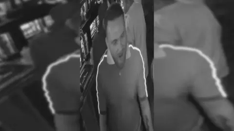 A black and white CCTV image of a man with short hair and a beard, wearing a collared t shirt.