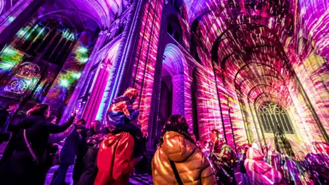 Luya A light installation, with vivid purple and yellow colours, is lighting up the inside of a cathedral building. There are people in the foreground, who are also lit up by the display, with some taking photos on their phones.