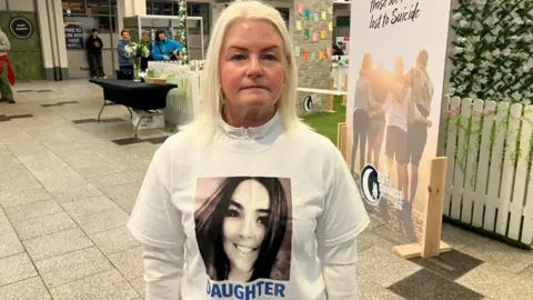 Colette Madden, with blond hair and white shirt. The shirt has an image of a woman and blue text under it saying the word daughter.