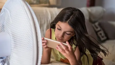 A girl looking at a mobile phone while lying on her tummy in front of a fan