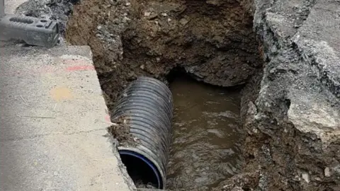 A hole in the ground with a black pipe in. Around the hole is grey concrete.