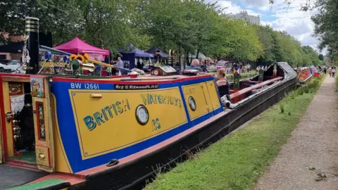 Lichfield & Hatherton Canals Restoration Trust A brightly decorated narrowboat moored along a canal towpath during a waterside event, with market stalls, visitors and trees lining the bank behind it.