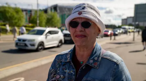 A woman wearing a white hate, sunglasses and denim jacket on a busy street. She has Heart of Midlothian badges on her jacket and hat.