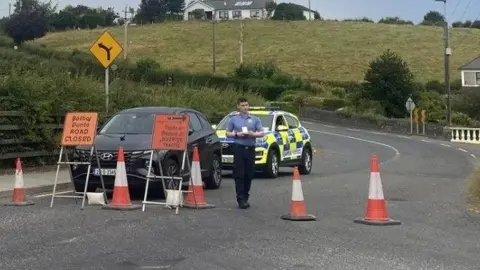 A yellow and blue police keep parked behind a black unmarked jeep. There are red cones closing the road and a police officer in uniform walking through them.
