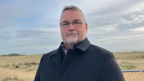 Henry Godfrey Evans/BBC Mark Stephenson wearing a black coat smiling at the camera. He is wearing small frame glasses and a black coat. Behind him is a beach.